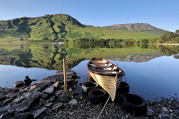 Small boat on the shore of Ballinakill Bay, sitting on tyres, with mountains reflected in the water. wp-image-1115