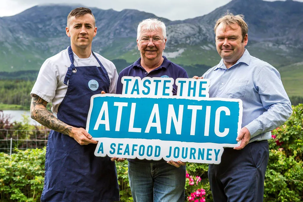 David Keane and 2 chefs hold a 'Taste The Atlantic' sign at the Oyster Farm in Ballinakill Bay. wp-image-1386